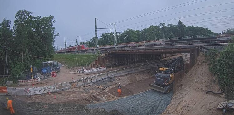 Eine Baustelle an einer Eisenbahnüberführung. Ein roter fährt Zug darüber.