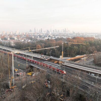 Eine Luftaufnahme der Arbeiten an der Golfstraße: Im Hintergrund steht die Skyline von Frankfurt, im Vordergrund fährt ein Zug über eine Brücke, links und rechts davon stehen zwei Baukräne.
