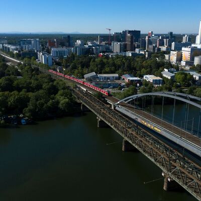 Ein roter Zug fährt auf eine Eisenbahnbrücke zu, darunter ein Fluss.