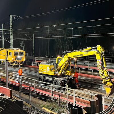 Ein gelber Bagger im Einsatz, im Hintergrund die Gleisstopfmaschine.