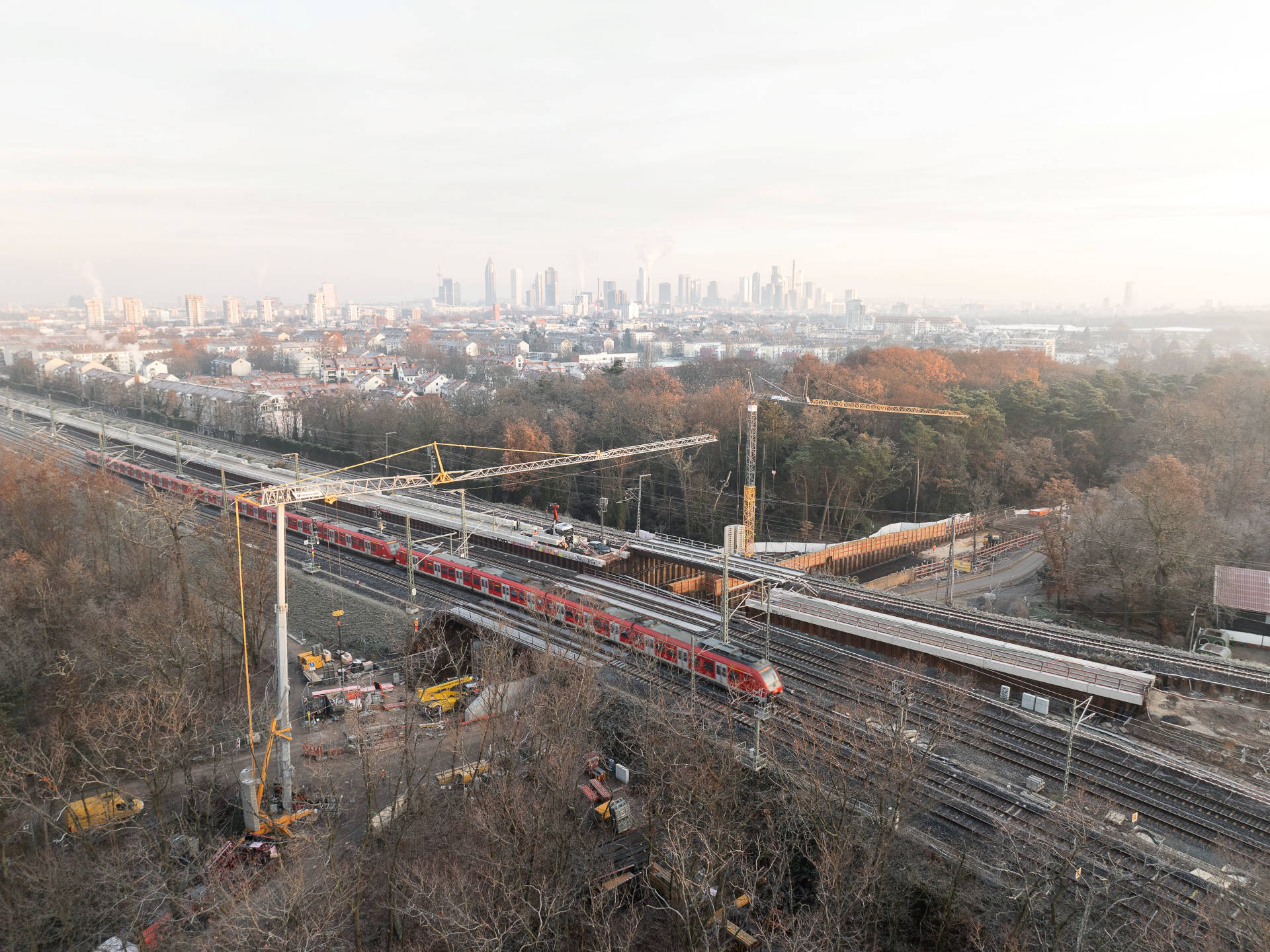 Eine Luftaufnahme der Arbeiten an der Golfstraße: Im Hintergrund steht die Skyline von Frankfurt, im Vordergrund fährt ein Zug über eine Brücke, links und rechts davon stehen zwei Baukräne.