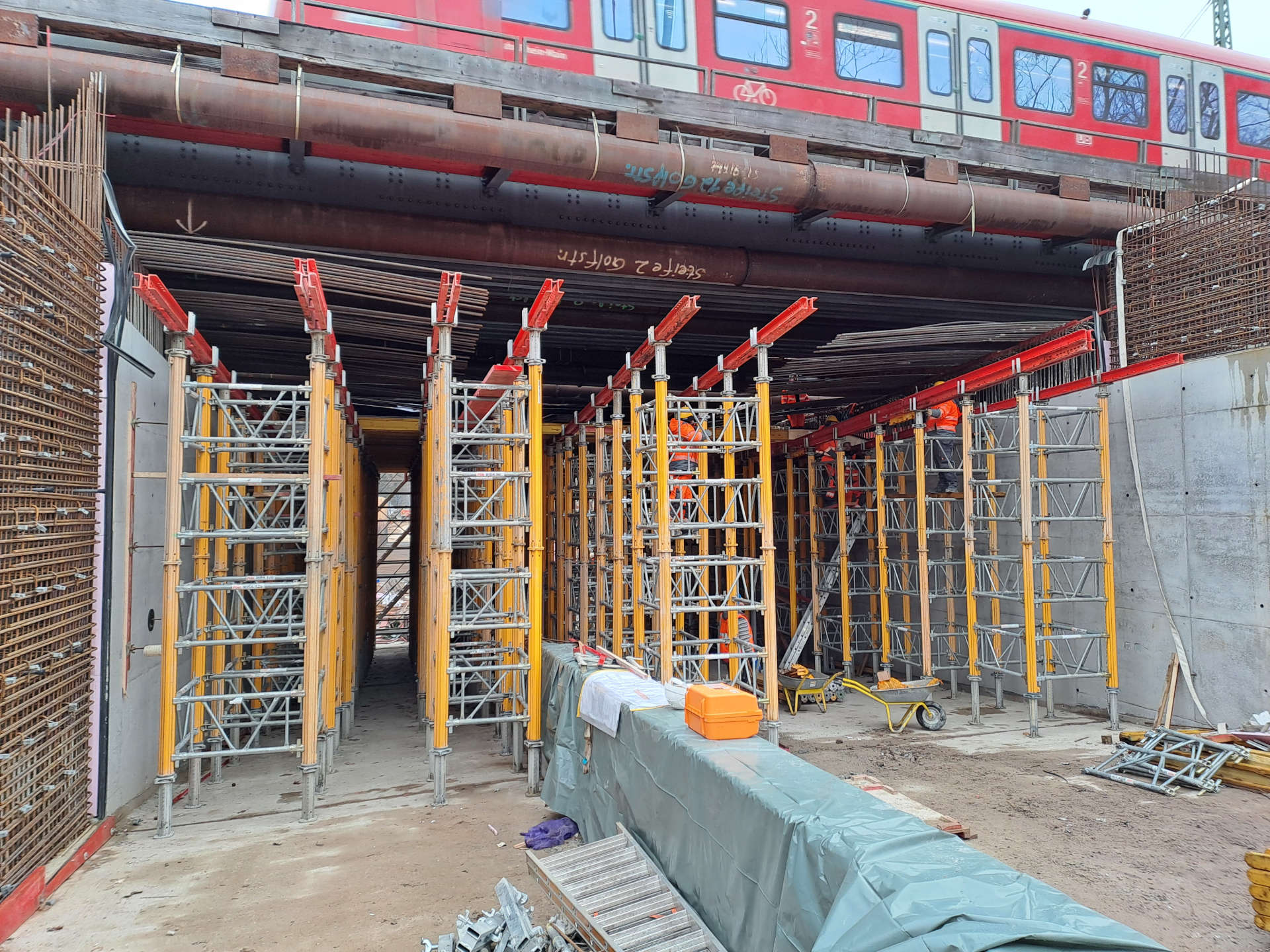 Schalungs- und Bewehrungsarbeiten an der Eisenbahnbrücke an der Golfstraße. Über die Brücke fährt oben ein Zug.